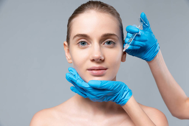 Portrait de jeune fille faisant des injections de cosmétologie sur les joues isolées sur fond de studio gris - Photo, image