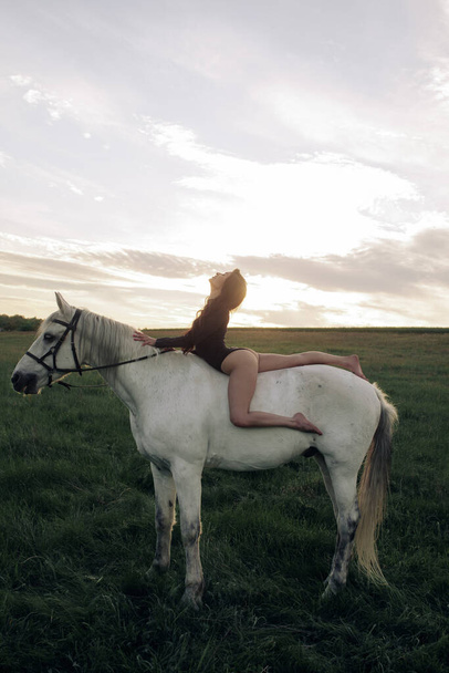 Genç kadın gün batımında beyaz at sırtında çayırda oturuyor.. - Fotoğraf, Görsel