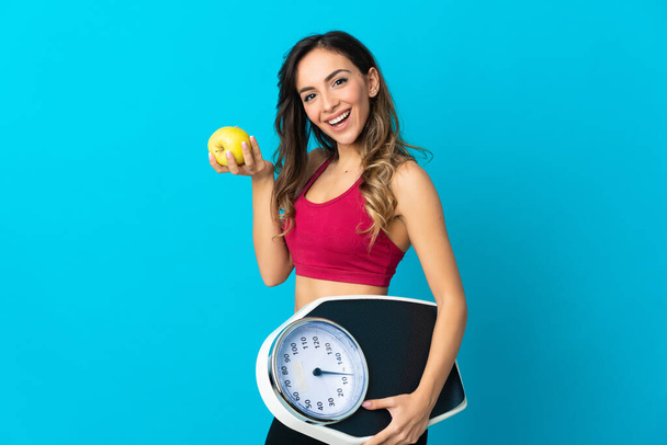 Young caucasian woman holding a scale isolated on blue background with weighing machine and with an apple - Fotoğraf, Görsel