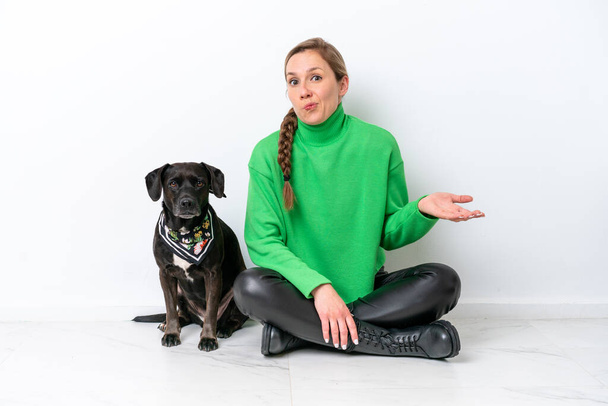 Young caucasian woman sitting on the floor with his puppy isolated on white background making doubts gesture while lifting the shoulders - Photo, Image