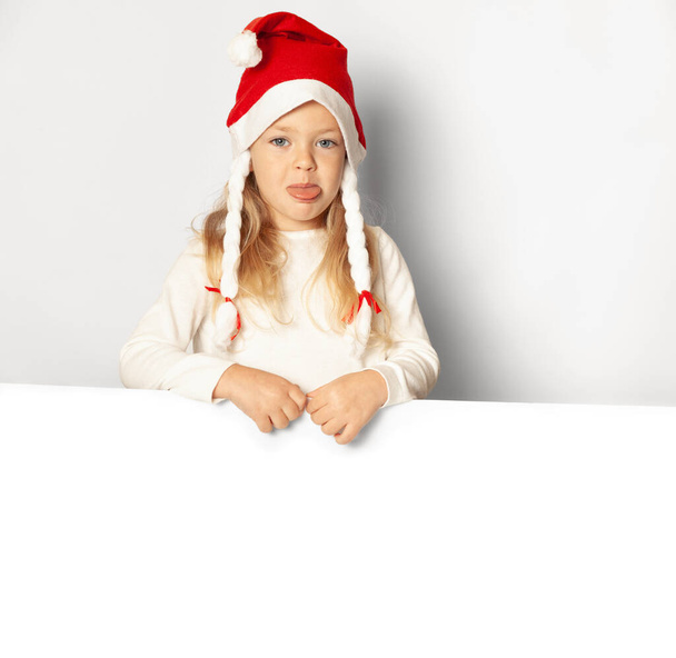 Portrait of little pretty girl upset in Santa hat sticks out tongue with sale (advertising) empty blank. Holding Xmas cardboard on white background. Happy New Year. - Foto, immagini