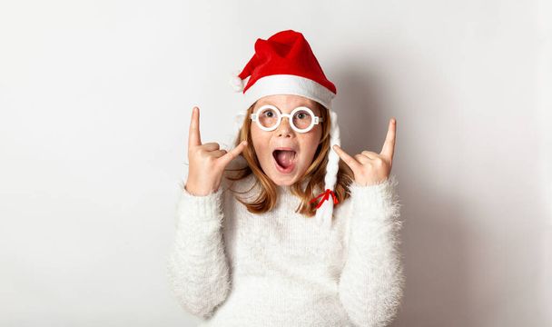 The beginning of the New Year's party, a girl in toy glasses and a Santa hat shows rock and roll with her fingers. - Photo, image