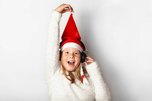 New Year and Christmas party, a girl in a Santa's Christmas hat listens to music on headphones, smiles and pulls a hat, isolated on a white background. - Foto, Imagem