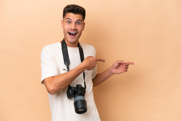 Nuori valokuvaaja valkoihoinen mies eristetty beige tausta yllättynyt ja osoittaa puolella - Valokuva, kuva