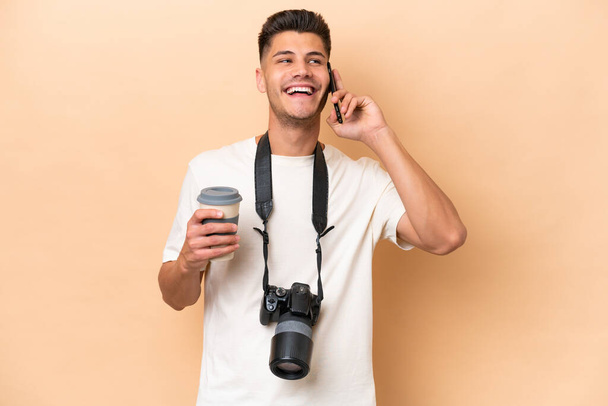 Young photographer caucasian man isolated on beige background holding coffee to take away and a mobile - Photo, Image