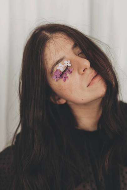 Beautiful sensual woman with lilac petals around closed eyes. Moody portrait of young brunette with flowers on face indoors. Skin care and natural cosmetics concept. Femininity Self care - Photo, Image