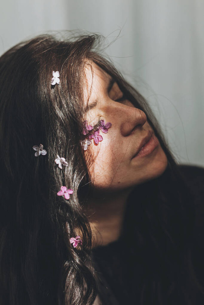 Beautiful sensual woman with lilac petals on face and hair in sunlight. Femininity. Moody portrait of young brunette with flowers on face in light and shadow. Skin care and self care. Femininity - Fotografie, Obrázek
