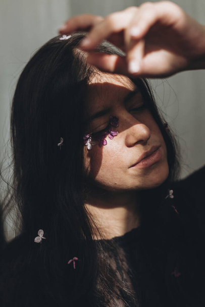 Beautiful sensual woman with lilac petals on face and hair posing in sunlight. Femininity. Moody portrait of young brunette with flowers on face in light and shadow. Skin care and self care - Φωτογραφία, εικόνα