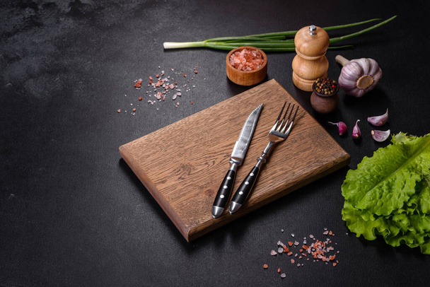 Вилка, нож, специи и травы, режущая доска на темном бетонном фоне. Кулинария дома - Фото, изображение