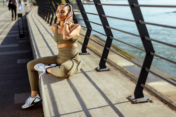 Pretty young woman with earphones takes a break after running in the urban area - 写真・画像