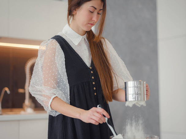 DIY home cooking concept. brunette woman sifting flour for cake through sieve, salting dough. Young woman smiling, loves to cook in beautiful modern kitchen. - Zdjęcie, obraz
