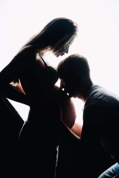 El marido besa el estómago de las mujeres embarazadas. Siluetas oscuras sobre fondo blanco. Foto del estudio. Foto de alta calidad - Foto, imagen