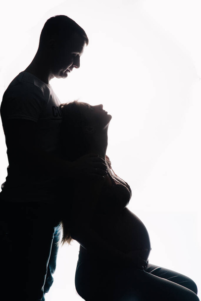 The silhouette of a man in a T-shirt and a pregnant woman in beautiful underwear on a white background. Studio photo. High quality photo - Foto, imagen