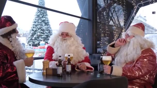 A group of Santa Clauses wish Merry Christmas and drink a glass of light beer. Three people in Santa costumes are talking at a Christmas party. - Video