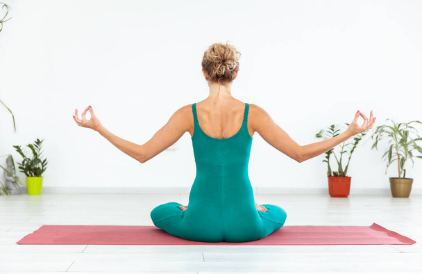Young yogi woman meditates while sitting on mat in yoga studio. Back view - 写真・画像