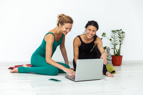 Duas amigas yogi estão sorrindo e usando um laptop enquanto estão sentadas no tapete no estúdio de ioga. Práticas de ioga online - Foto, Imagem