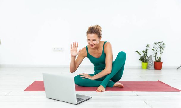 Улыбающаяся женщина-йог приветствует своих учеников, глядя в экран ноутбука, сидя на коврике в студии йоги. Уроки йоги онлайн - Фото, изображение