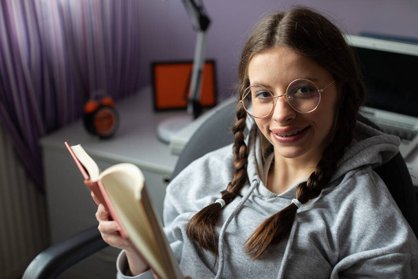 A cheerful girl is happily reading her favorite book. The brunette has her glasses on and her hair is styled in two braids. She loves reading books. - Photo, image
