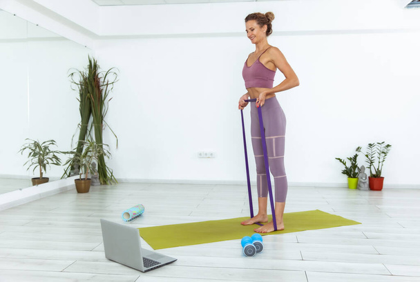 Mulher fitness assistindo aula on-line no laptop e exercício com uma faixa de borracha fitness dentro de casa - Foto, Imagem