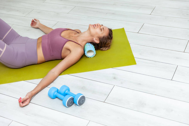 jonge vrouw ontspant de spieren van de nek door ze te masseren met een massage roller terwijl liggend op de mat - Foto, afbeelding