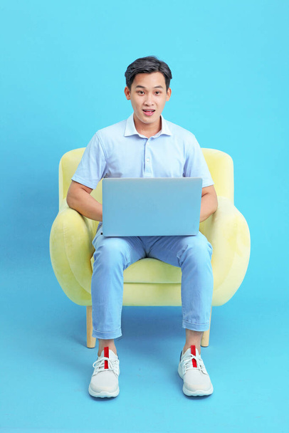 Asian handsome and cheerful young man working on laptop and sitting on sofa, isolated on background - Foto, Imagem