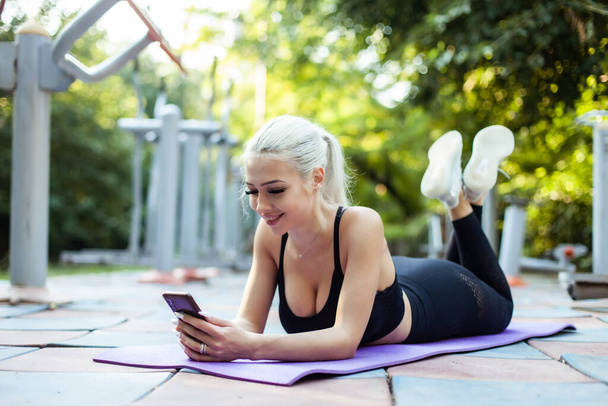 Fitness nő használ okostelefon, miközben fekszik a szőnyegen a sportpályán - Fotó, kép