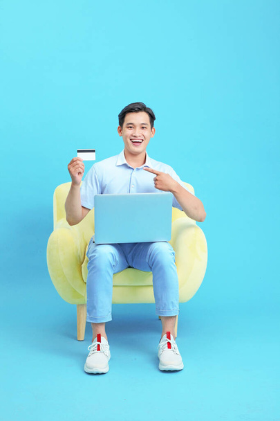 Asian young handsome guy, sitting on sofa, using credit or debit card to make online payment on laptop computer, isolated on blue background - Foto, Imagem
