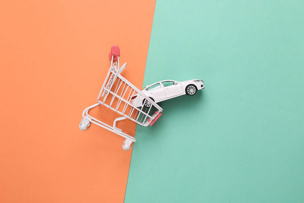 Buying a car concept. Shopping cart with toy car model on pink blue background. Top view - Photo, Image