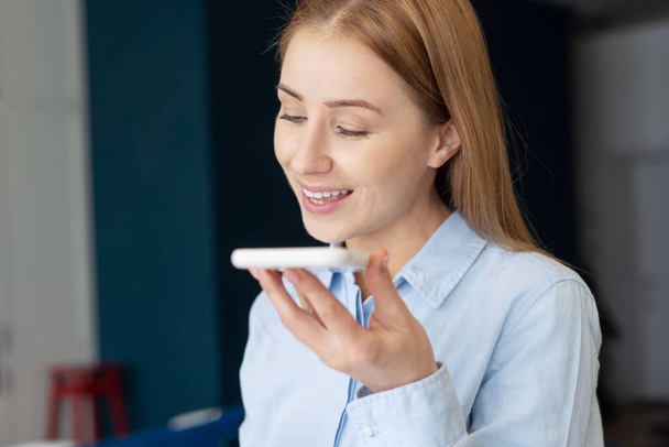 Πορτρέτο του χαμογελαστού γυναίκα καταγραφή ηχητικό μήνυμα στο smartphone, χρήση εικονική βοηθός φωνή στη σύγχρονη gadget. Νεαρή γυναίκα μιλάει σε ανοιχτή ακρόαση με τηλεφωνικό κέντρο στο κινητό της - Φωτογραφία, εικόνα