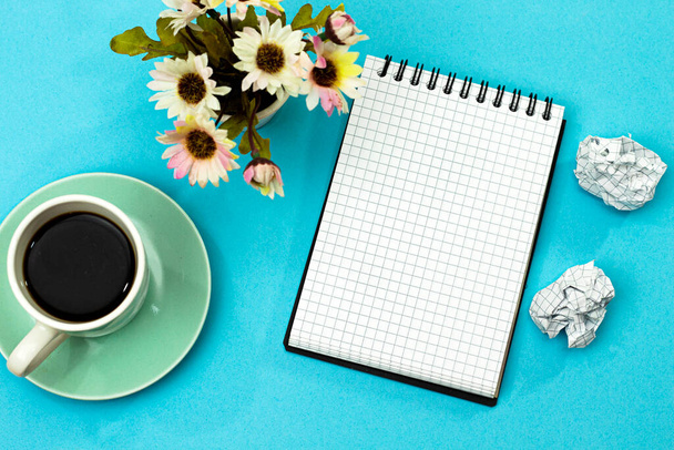 επίπεδη lay επιφάνεια εργασίας, συντριβή χαρτιά, γλάστρα λουλούδι και φλιτζάνι καφέ γάλα με σημειωματάριο. σε μπλε φόντο - Φωτογραφία, εικόνα
