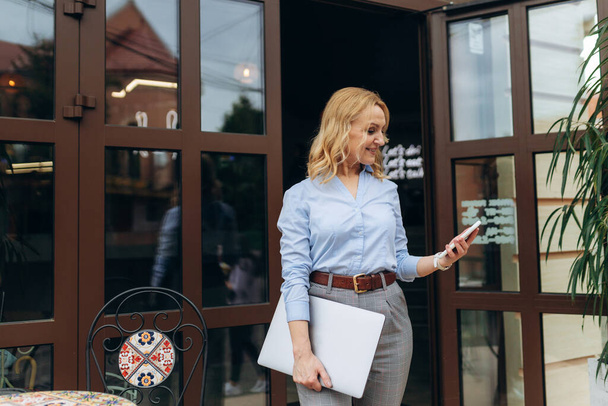 Portret szczęśliwej dojrzałej bizneswoman korzystającej z telefonu stojącej na chodniku kawiarni - Zdjęcie, obraz