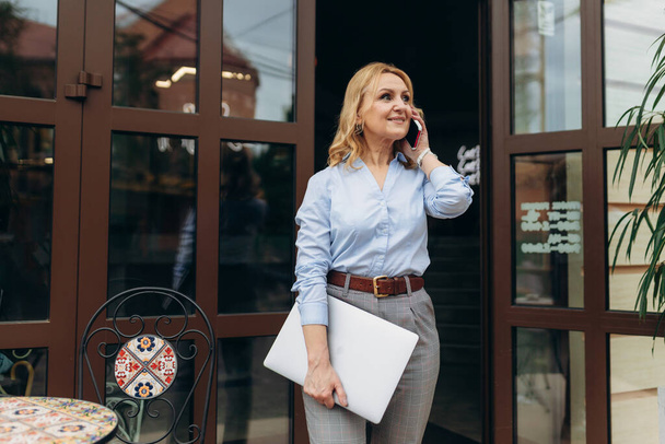 Портрет счастливой зрелой бизнесвумен с помощью телефона, стоя на тротуаре кафе - Фото, изображение