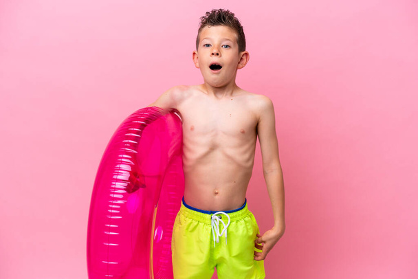 Pequeño chico caucásico sosteniendo una rosquilla inflable aislada sobre fondo rosa con expresión facial sorpresa - Foto, imagen
