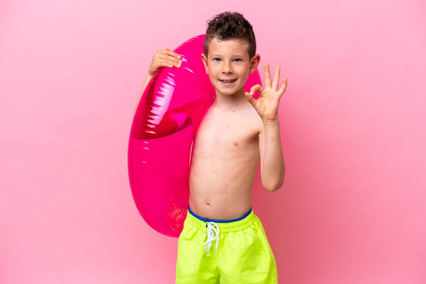 Pequeño chico caucásico sosteniendo una rosquilla inflable aislada sobre fondo rosa mostrando signo de ok con los dedos - Foto, imagen