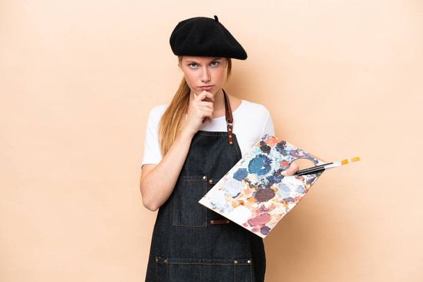 Молода художниця біла жінка тримає палітру ізольовано на бежевому фоні мислення
 - Фото, зображення