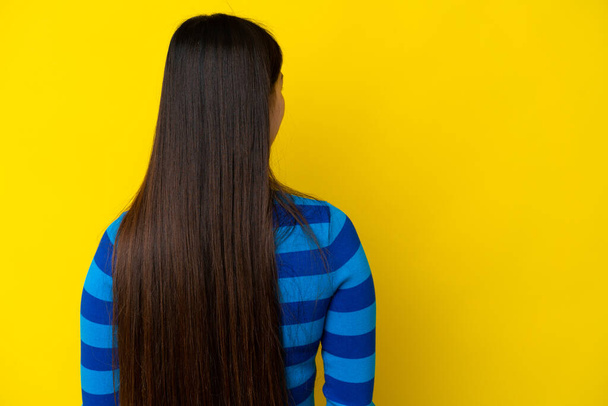 Joven mujer china aislada sobre fondo amarillo en posición trasera y mirando hacia el lado - Foto, imagen