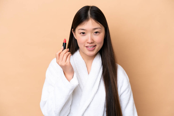 Young Chinese woman isolated on beige background holding red lipstick - Foto, imagen