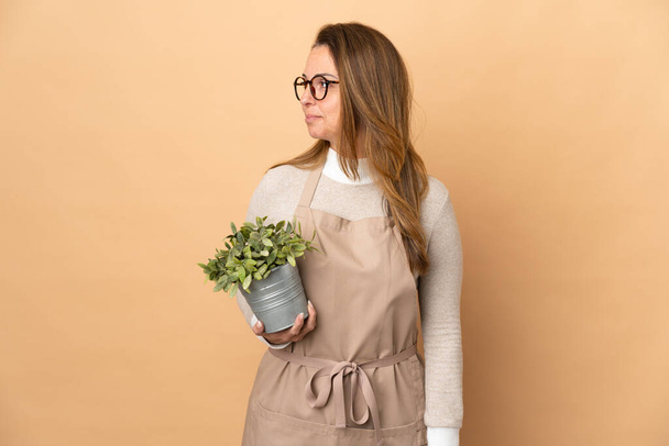 Mujer jardinero de mediana edad sosteniendo una planta aislada sobre fondo beige mirando hacia un lado - Foto, Imagen