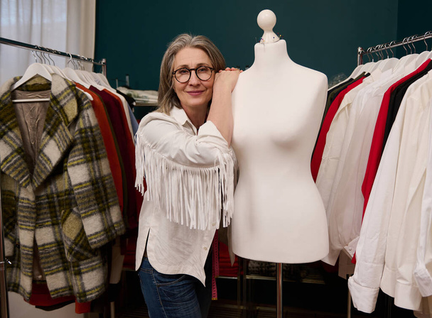 Charming mature female fashion designer posing with a tailoring mannequin, enjoying her creative work in fashion clothing design atelier - 写真・画像