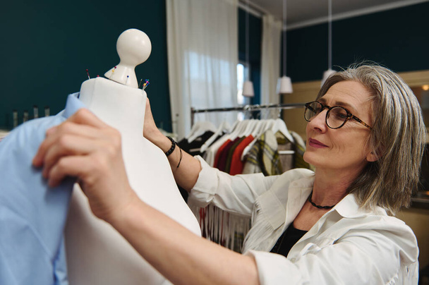 Mature Caucasian woman fashion designer tailor, seamstress tries on a blue shirt from a new collection on a mannequin in her clothing design and tailoring workshop - 写真・画像
