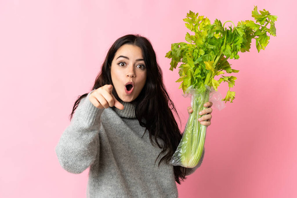 Young caucasian woman holding a celery isolated on blue background surprised and pointing front - Photo, Image