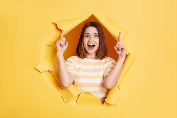 Portrait of surprised amazed woman stands in torn paper hole, pointing fingers up, presenting copyspace, looking through breakthrough of yellow background. - Foto, Imagen