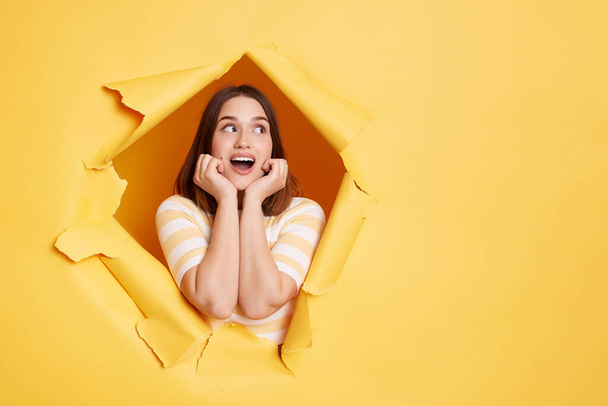 Indoor shot of surprised amazed brunette woman stands in torn paper hole, looks away with open mouth, keeps hands under chin, looking through breakthrough of yellow background. - Foto, Imagen