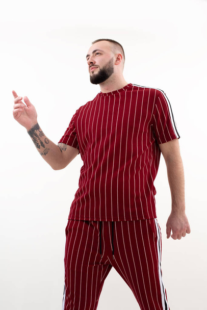 Vertical portrait of brunet tattooed confident man posing in studio on white background. Pointing forefinger. Full length male gesturing by wearing informal red sportswear. Modern successful man - Photo, image
