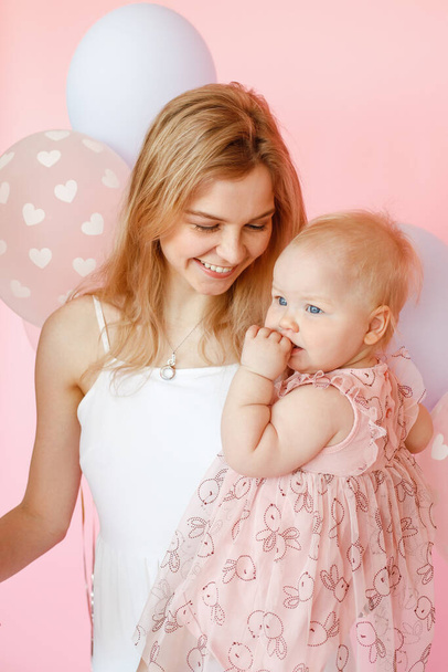 Portrait closeup of young smiling mother holding cute baby girl in holiday dress on pink background. First birthday party, pink decor and balloons. Happy moments and family holiday celebration. - Photo, Image