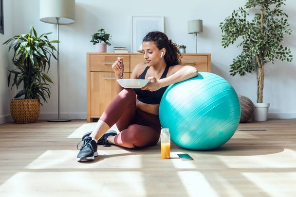 Tiro de mujer joven y deportiva comiendo sano mientras escucha música sentada en el suelo en casa. - Foto, Imagen