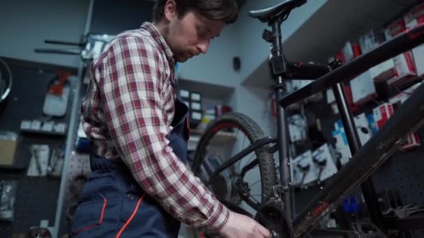 Zmęczony mechanik rowerowy pracujący w sklepie rowerowym naprawiający rowery klientów. Naprawiacz naprawia dolny wspornik na rowerze górskim. Wymiana systemu łącznika. Koła łańcuchowe i korby - Materiał filmowy, wideo