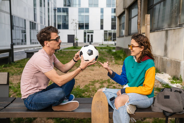 Couple woman and young caucasian man modern happy adult male and female boyfriend and girlfriend smile sitting in front of stadium holding soccer ball outdoor in day football game concept copy space - Фото, изображение