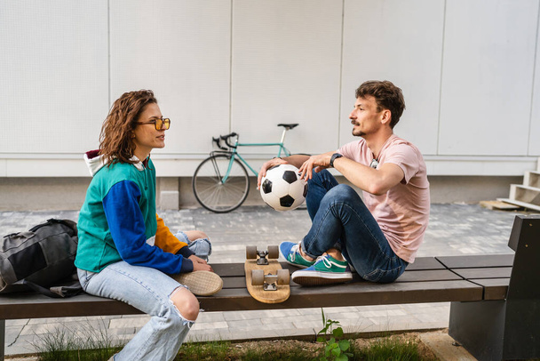 Couple woman and young caucasian man modern happy adult male and female boyfriend and girlfriend smile sitting in front of stadium holding soccer ball outdoor in day football game concept copy space - 写真・画像
