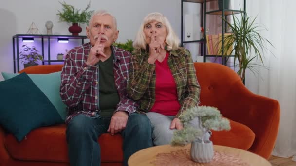 Stil zijn, alsjeblieft. Oudere familie paar grootouders man vrouw drukt index vinger op de lippen maakt stilte gebaar teken vertelt geen geheim. volwassen grootmoeder grootvader op bank thuis - Video